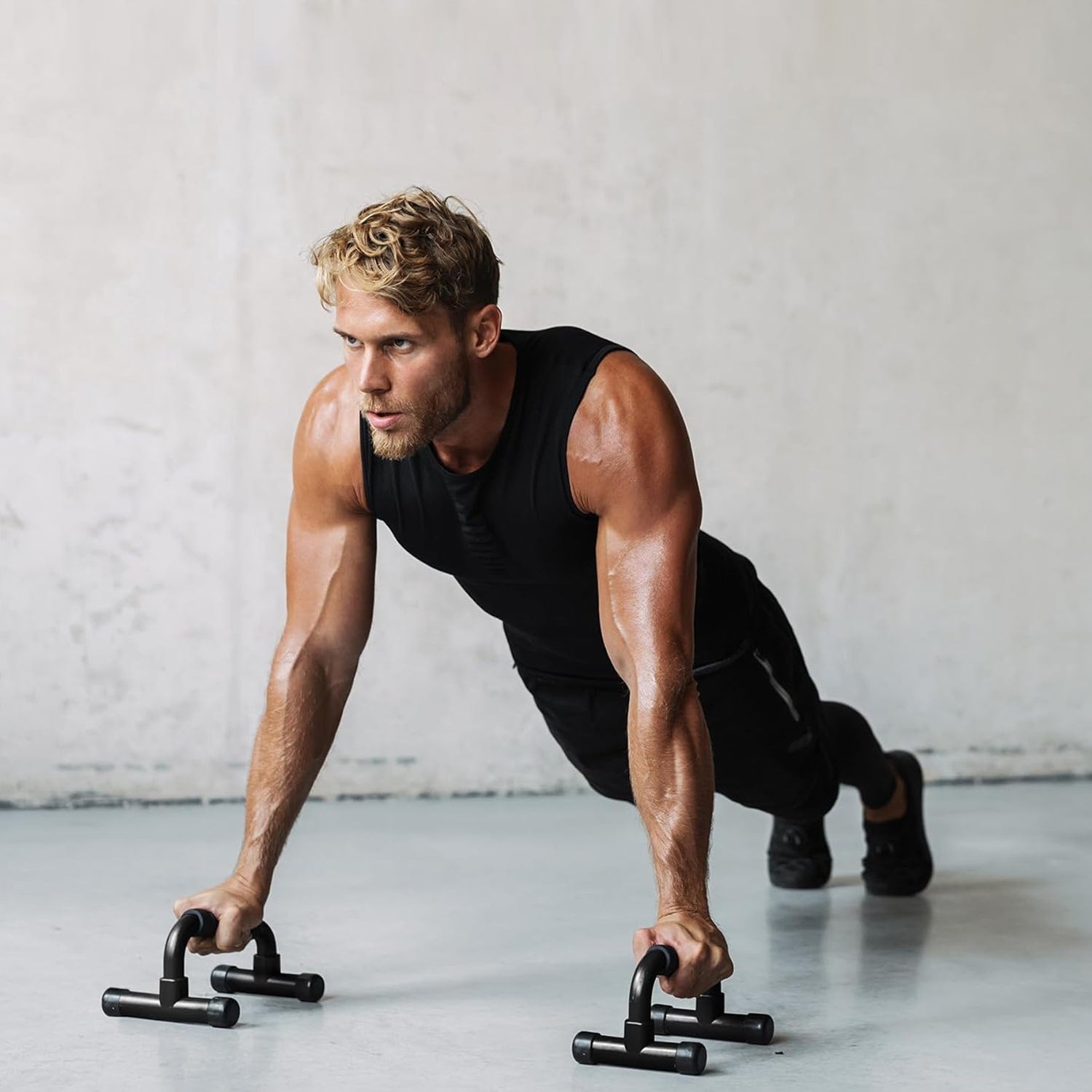 A fit, muscular man in a black sleeveless shirt and workout pants is performing push-ups using push-up bars. His arms and shoulders are visibly engaged, showing strength and focus. The background features a minimalistic, neutral-toned wall, emphasizing the intensity of his workout. Firstfit