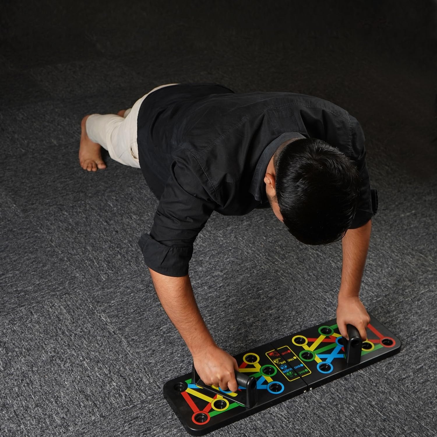 A man in a black shirt and beige pants is performing a push-up using a colorful push-up board on a dark carpeted floor. The push-up board has multiple color-coded positions for targeting different muscle groups. His posture and focus suggest an engaged workout session. Firstfit